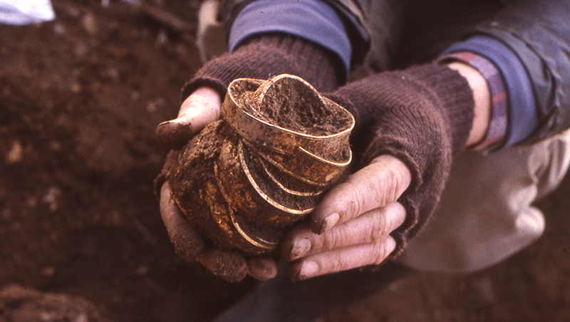 a person holding the gold bracelets in gloved hands and lifting them from the trench.